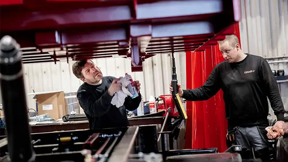 Zwei Arbeiter in schwarzen DISAB Uniformen führen Wartungsarbeiten an einer roten Industrieeinheit durch, wobei sie Werkzeuge und Reinigungsmittel in einer gut ausgestatteten Werkstatt verwenden.