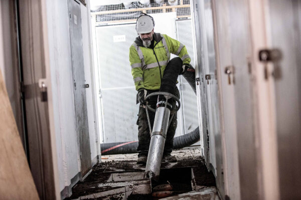 Bediener in Schutzkleidung, der einen großen Saugschlauch einer DISAB-Einheit in einem Gebäude verwendet, wobei Sicherheit, Ergonomie und Materialhandhabung in beengten Industrieräumen betont werden.